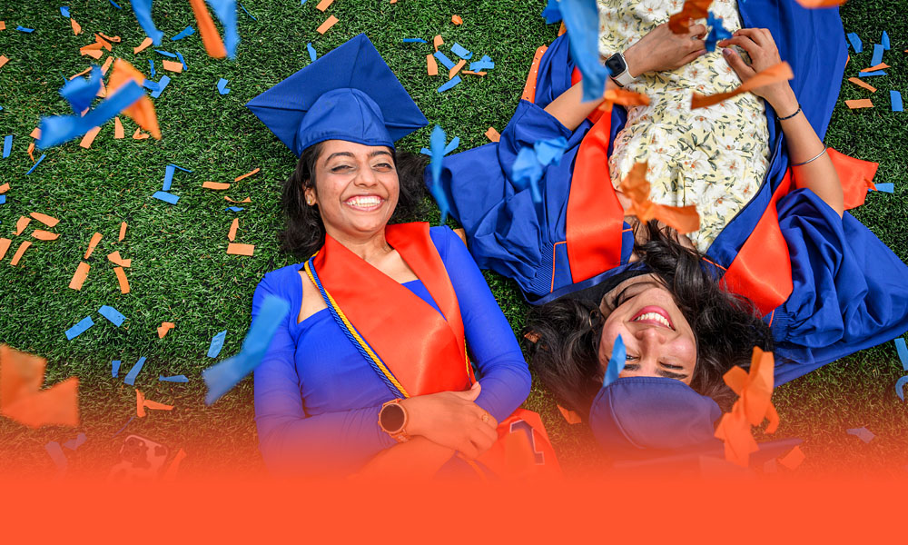 two smiling graduating students laying on orange and blue confetti grass at Memorial Stadium