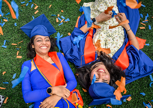 overhead of two smiling graduating students laying on the orange and blue confetti grass at Memorial Stadium